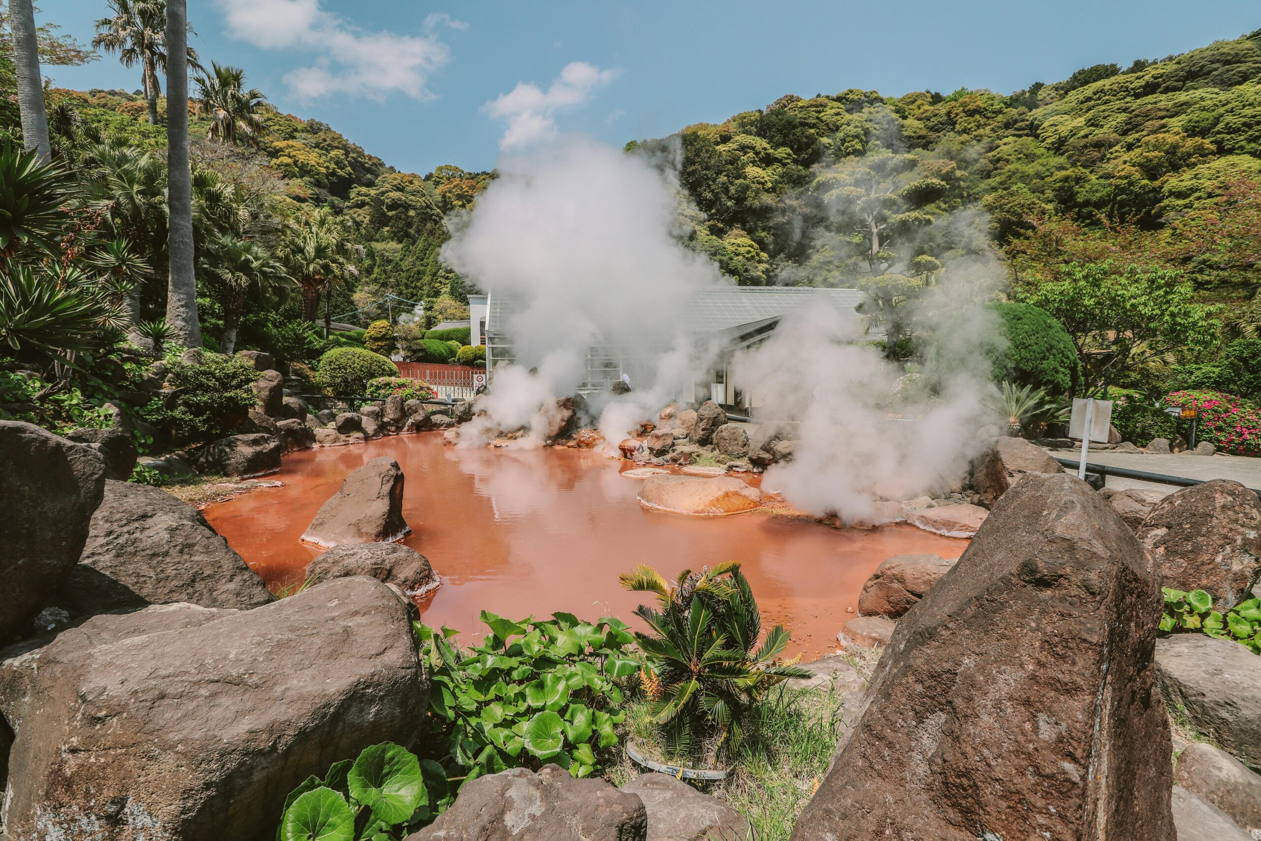 Explore a vibrant hot spring surrounded by lush greenery with rising sulfur smoke under a clear blue sky.